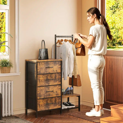 A woman stands beside a rustic three-drawer chest and clothing rack in a bright room with wood floors and windows. The chest has a weathered brown wood top and black metal frame, with a black tote bag resting on it.
