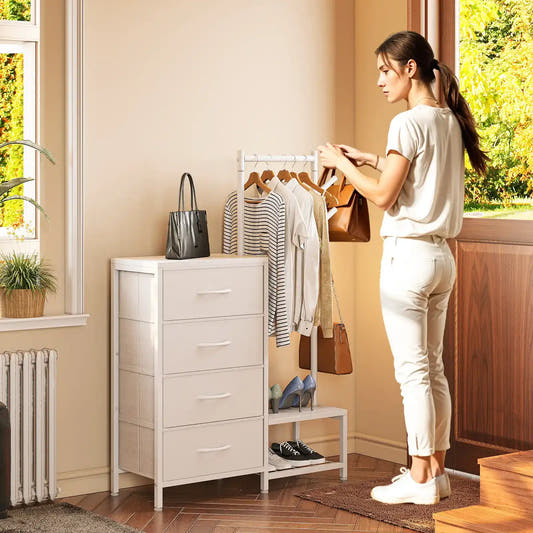 A woman stands beside a white 4-drawer storage cabinet in a bright room, with a clothing rack, bags, and shoes nearby. The cabinet is the likely product being shown, photographed in a real interior setting with a person present, so subject_type is Lifestyle.