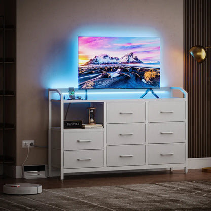 A white modern TV stand cabinet with multiple drawers and open shelving, holding a television and small decor items, in a styled living room setting with ambient blue lighting and a robot vacuum nearby.