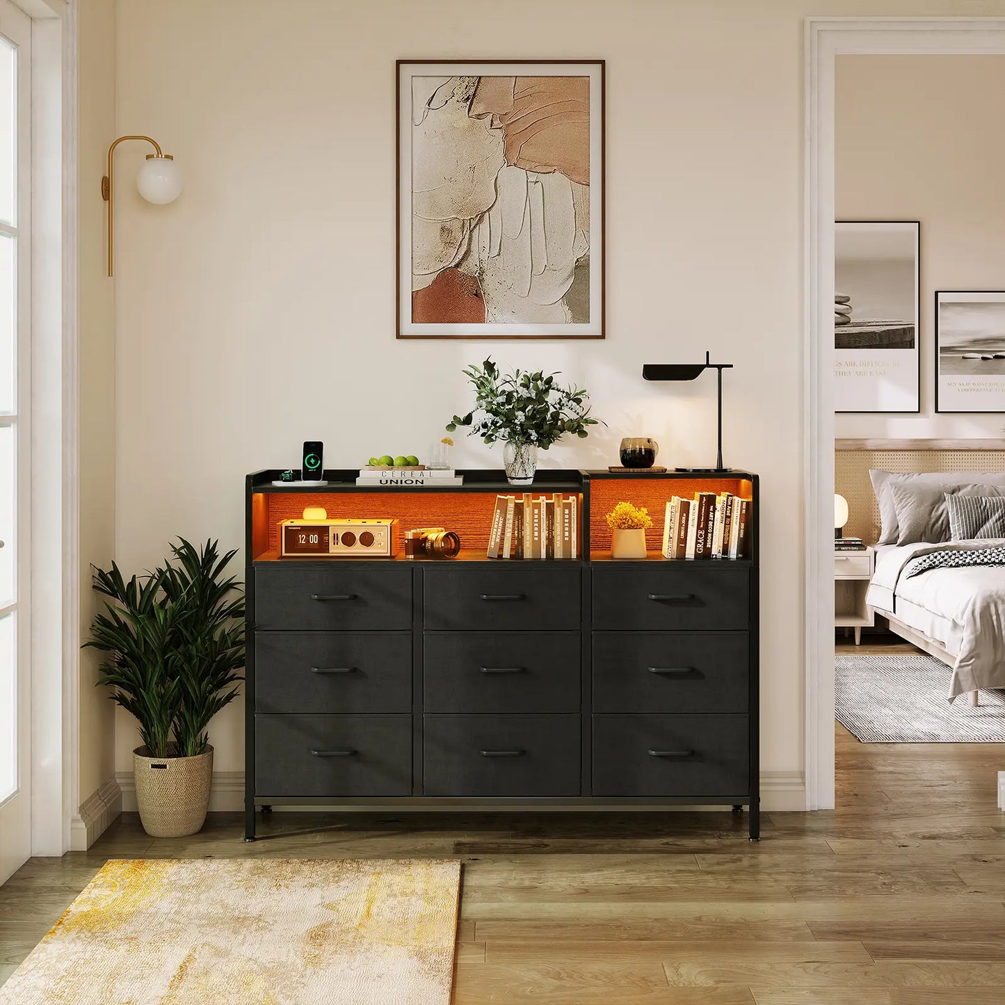 A modern black wooden dresser with six drawers and an open upper shelf illuminated by warm orange lighting, styled in a bedroom entryway with books, a lamp, and a plant.