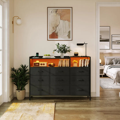 A modern black wooden dresser with six drawers and an open upper shelf illuminated by warm orange lighting, styled in a bedroom entryway with books, a lamp, and a plant.
