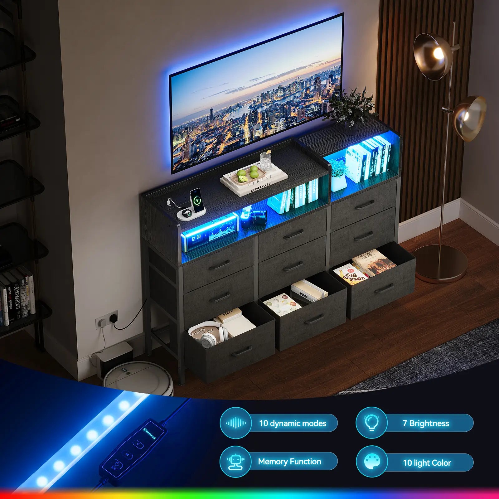 A dark gray modern TV stand with built-in LED lighting, shown in a styled living room setting with a TV, floor lamp, and shelving. The stand features multiple drawers, open shelves, and illuminated compartments displaying books and accessories.