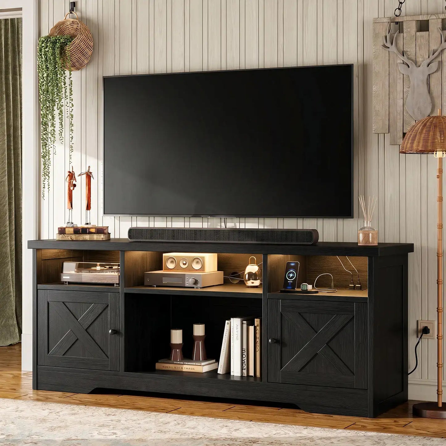 A black wooden TV stand/media console with two cabinet doors, open shelving, and a soundbar, shown in a styled living room setting with a large black television mounted above it.