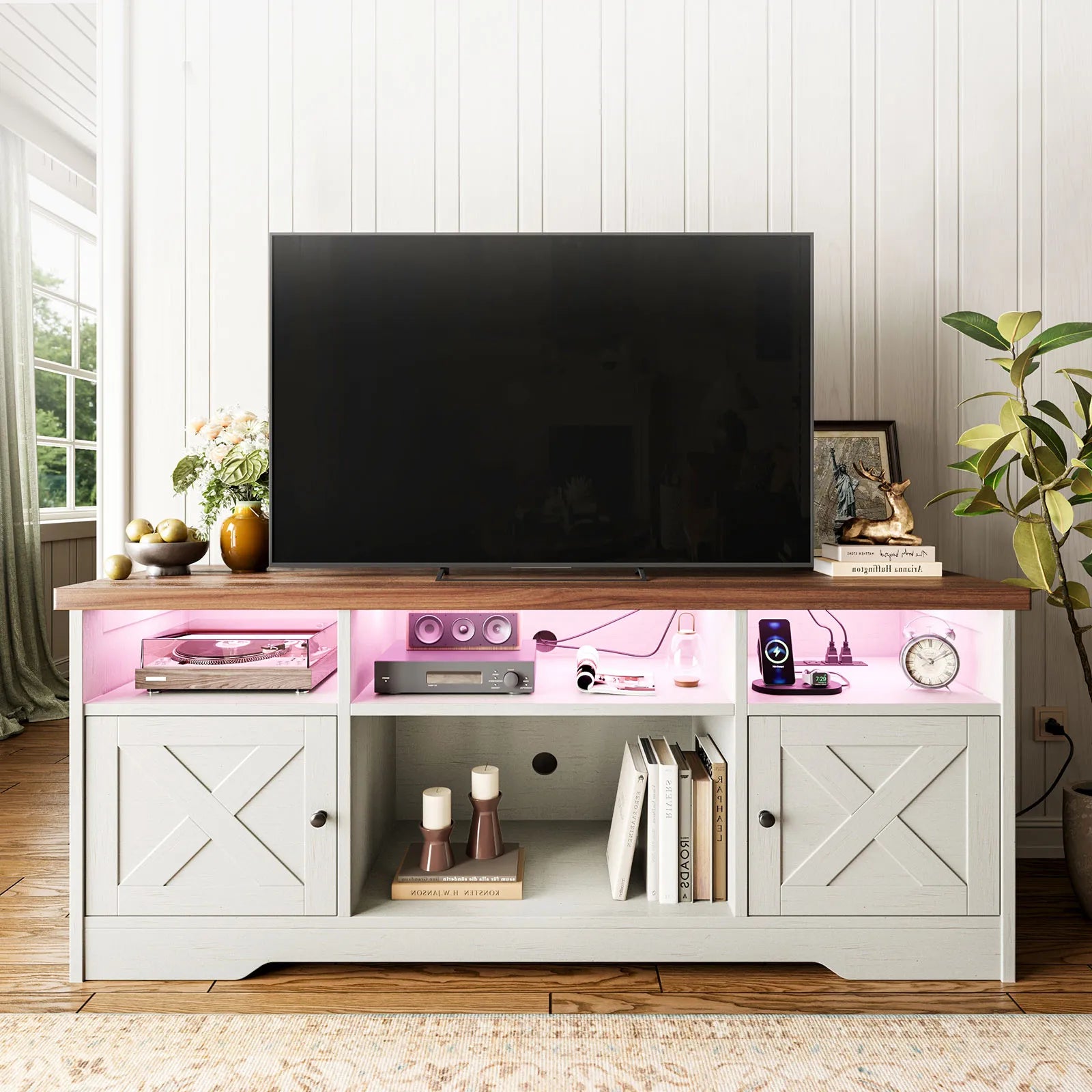 A light gray TV stand with a dark wood top, featuring X-brace cabinet doors, open shelving, and pink illuminated interior lighting, shown in a styled living room setting with a television and home decor items.